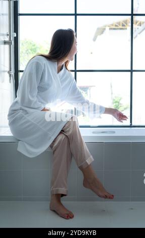 Femme assise sur la baignoire portant un peignoir blanc dans la salle de bain moderne à la maison avec des feuilles de rose pour rehausser le parfum du bain Banque D'Images