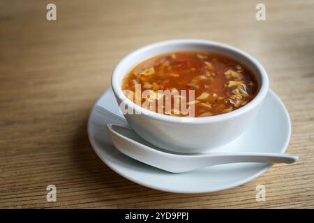 Soupe chinoise chaude et aigre avec des œufs battus, de la viande et des légumes dans un bouillon de poulet, bol blanc avec une cuillère en porcelaine sur une table en bois dans un AS Banque D'Images