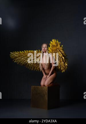 Modèle pose gracieusement avec des ailes dorées sur un piédestal dans un cadre de studio sombre Banque D'Images