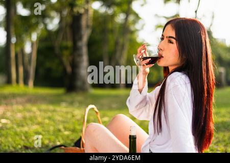 gros plan d'une fille de latina aux cheveux rougeâtres buvant un verre de vin tout en étant assise sur le sol dans le parc. Banque D'Images