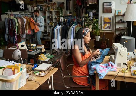 Jeune femme en tenue décontractée assise par table avec machine à coudre électrique et veste en denim décorative avec des perles contre le client Banque D'Images