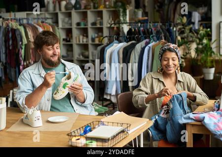 Heureux jeune homme et femme assis près de tables dans la friperie et upcycling utilisé veste en denim et chaussures croisées Banque D'Images