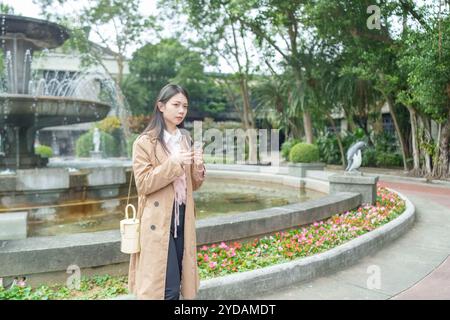 Une taïwanaise aux cheveux longs dans la vingtaine portant un trench-coat beige utilise un smartphone autour d'une fontaine dans un parc dans le district de Xinyi, Taipei City, Banque D'Images