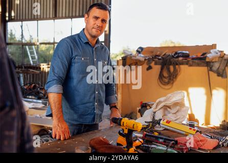Un jeune homme souriant en vêtements décontractés se tient à une table avec des outils de construction. Banque D'Images