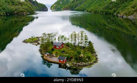 Une petite île avec deux cabanes rouges nichées parmi les arbres verts est entourée d'eaux calmes et réfléchissantes. Lovrafjorden, Norvège Banque D'Images