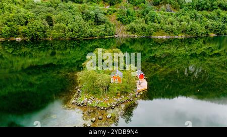 Petite île verte avec deux cabanes rouges nichées parmi les arbres dans un fjord norvégien isolé. l'eau calme reflète l'environnement luxuriant, créant un ser Banque D'Images