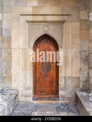 Une porte en bois magnifiquement conçue se dresse à l'entrée d'un bâtiment historique en pierre, présentant des sculptures complexes et une arche unique. Les murs environnants sont faits de grosses pierres vieillies. Banque D'Images