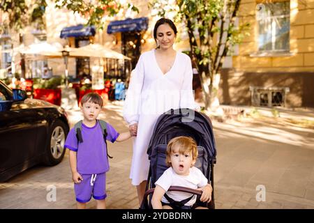 Femme enceinte en robe blanche marche avec ses deux enfants autour de la ville. Maman avec poussette mène son enfant aîné à la main sur stree Banque D'Images