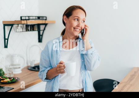 Jeune femme souriante attrayante avec une tasse de café dans la cuisine parlant au téléphone. Banque D'Images