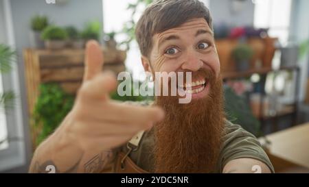 Homme barbu joyeux pointant à l'intérieur entouré de plantes dans un magasin de fleurs lumineux Banque D'Images