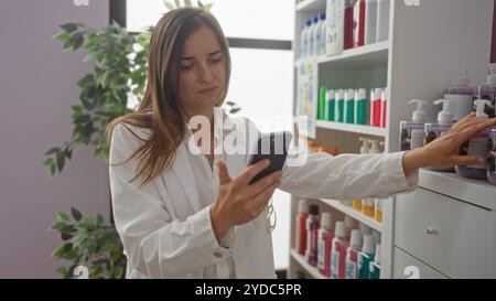 Une jeune femme en manteau blanc utilise son téléphone tout en sélectionnant des produits dans une pharmacie très éclairée. Banque D'Images