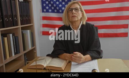 Femme juge portant des lunettes et une robe noire assise au bureau dans le bureau avec drapeau américain en arrière-plan et étagères à côté d'elle Banque D'Images