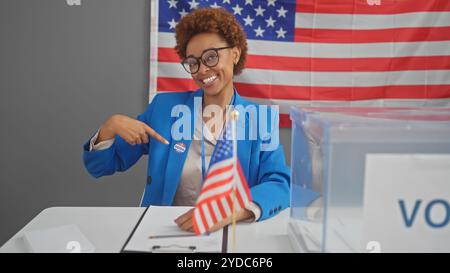 Femme afro-américaine souriante dans un blazer bleu pointe sur l'autocollant "j'ai voté", avec nous drapeau et urne. Banque D'Images