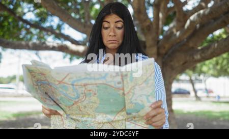 Une belle femme hispanique tient une carte et semble concernée dans un parc extérieur sous un grand arbre par une journée ensoleillée. Banque D'Images