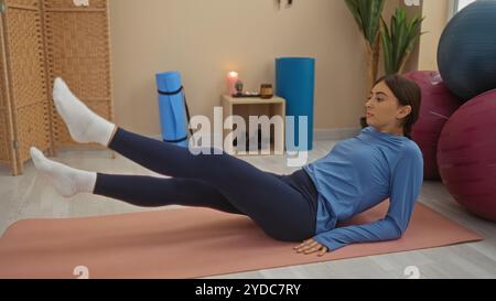 Femme exerçant sur un tapis dans un intérieur de gymnastique, effectuant des levées de jambe avec un bolster de fitness bleu et des ballons d'exercice en arrière-plan, mettant en valeur athlétique Banque D'Images