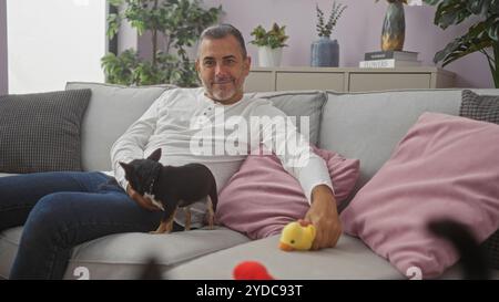 Homme hispanique d'âge moyen assis avec son chien chihuahua dans un salon confortable, entouré de coussins et de plantes décoratives, tenant un canard jouet. Banque D'Images