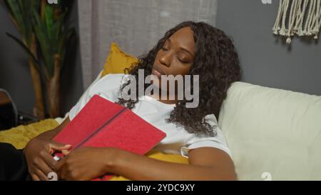 Une jeune femme afro-américaine aux cheveux bouclés dort paisiblement sur un lit dans une chambre intérieure confortable, serrant un livre rouge à sa poitrine. Banque D'Images