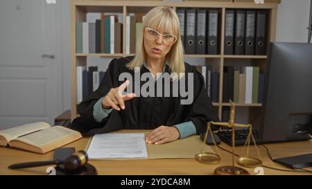 Une femme juge travaillant dans un bureau de salle d'audience s'est concentrée sur les documents juridiques, portant des lunettes et des robes de juge, avec un marteau et une balance de justice sur la ta Banque D'Images