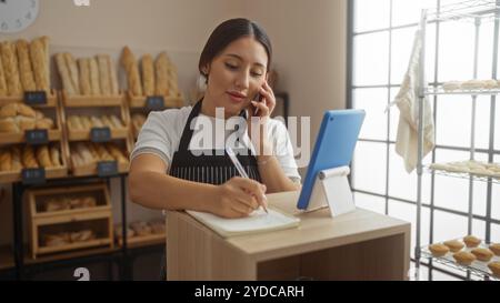 Jeune femme travaillant dans une boulangerie parlant au téléphone et prenant des notes avec du pain sur des étagères en arrière-plan et une tablette sur le comptoir Banque D'Images