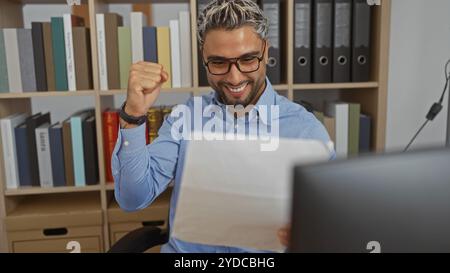 Jeune homme arabe dans un bureau lisant un document, souriant et faisant une pompe à poing, célébrant une réalisation avec des étagères en arrière-plan. Banque D'Images