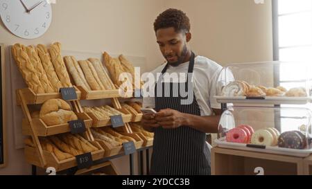 Jeune homme utilisant un smartphone dans une boulangerie avec du pain frais et des pâtisseries affichées à l'intérieur sur des étagères et des comptoirs Banque D'Images