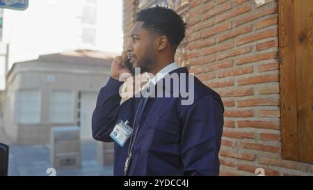 Homme afro-américain en vêtements de travail parlant sur un téléphone dans une rue de la ville avec un badge et une toile de fond de mur de briques. Banque D'Images