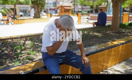 Un homme hispanique inquiet, aux cheveux gris, d'âge moyen, est assis dans un parc urbain, couvrant son visage avec sa main inquiète. Banque D'Images