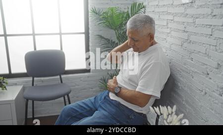 Un homme hispanique aux cheveux gris en chemise blanche est assis dans une salle d'attente intérieure lumineuse, tenant son cou dans l'inconfort à côté d'une chaise moderne et d'un pl vert Banque D'Images