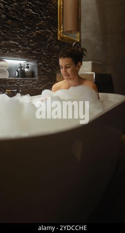Une femme souriante se détend dans un bain moussant dans un intérieur de salle de bain luxueux, exsudant la sérénité et le confort. Banque D'Images
