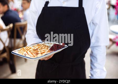 Gros plan des mains du serveur tenant le plateau avec du fromage à pâte molle frite et de la confiture. L'homme dans le tablier noir porte l'assiette avec le fromage mariné grillé traditionnel chypriote Banque D'Images