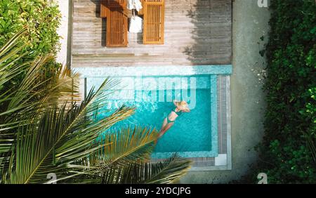 Une jeune femme en chapeau et maillot de bain se détache dans l'eau claire d'une piscine privée. Vue de dessus de la magnifique piscine avec chaises longues. La maison est surrou Banque D'Images