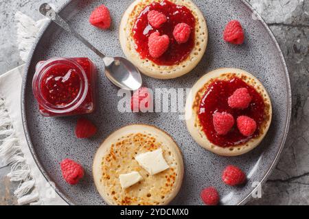 Des crumpets fraîchement grillés sur les plats de service avec du beurre, de la confiture et des framboises fraîches gros plan sur l'assiette sur la table. Vue horizontale de dessus Banque D'Images