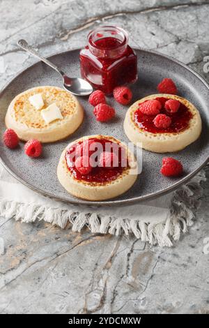 Crumpet est une crêpe ronde à base de pâte spongieuse servie avec du beurre, de la confiture et des framboises fraîches en gros plan dans une assiette sur une table. Vertical Banque D'Images