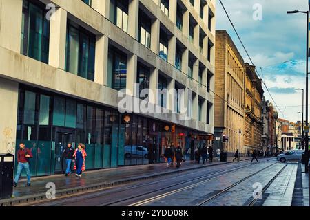 Une scène de rue avec un bâtiment moderne sur la gauche et des bâtiments plus anciens sur la droite. La rue est humide et a des voies de tramway dans le centre. Il y a du pape Banque D'Images