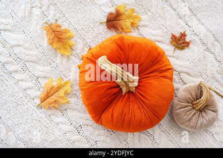 La grande citrouille jouet en tissu orange vif est placée sur un fond de couverture tricotée blanche confortable, accompagnée d'une citrouille plus petite et de feuilles d'automne dispersées Banque D'Images