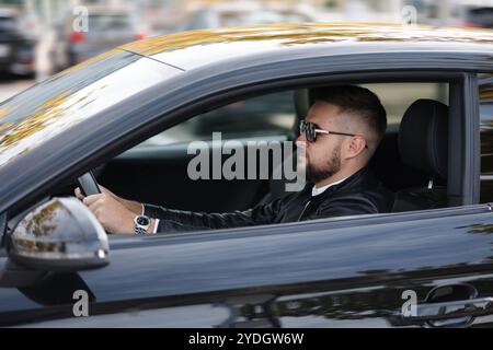 Succès en mouvement. Beau jeune homme en veste de cuir et lunettes de soleil conduisant une voiture. Vue latérale de l'extérieur Banque D'Images