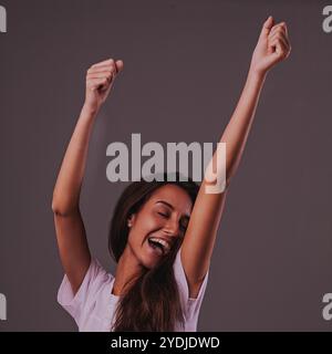 Jeune femme excitée en t-shirt blanc célèbre le succès avec les bras levés et les yeux fermés, montrant la joie sur fond gris Banque D'Images