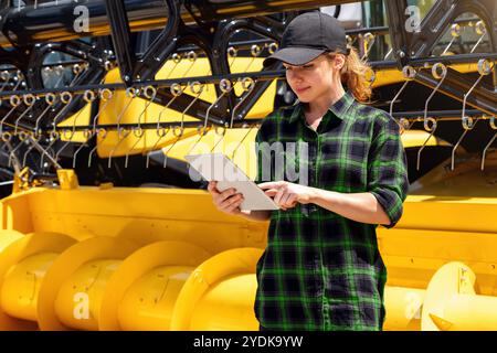Concessionnaire de matériel agricole. Femme dans une chemise décontractée verte se tient à côté d'une moissonneuse-batteuse jaune, travaillant sur une tablette numérique. Banque D'Images