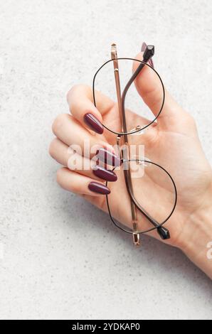 Une main avec de longs ongles Bordeaux tient gracieusement une paire de verres ronds au-dessus d'une surface texturée légère, éclairée par une lumière naturelle douce. Banque D'Images