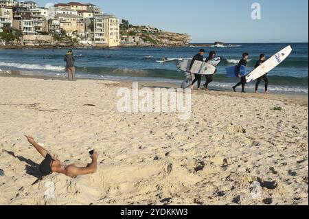 21/09/2018, Sydney, Nouvelle-Galles du Sud, Australie, Un jeune homme gît à moitié enterré dans le sable de Bondi Beach, tandis qu'un groupe de jeunes surfeurs marchent le long de la plage Banque D'Images