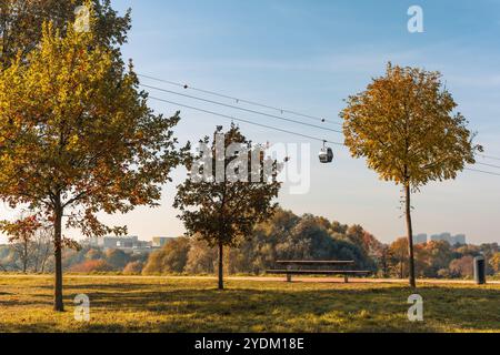 Un parc avec un banc et un arbre, un téléphérique suspendu en arrière-plan. Le ciel est bleu et les arbres sont pleins de feuilles Banque D'Images