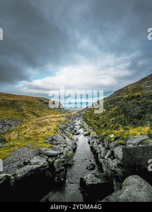 Un ruisseau étroit coule à travers une vallée, entourée de gros rochers et d'une petite quantité de végétation verte. Le ciel est nuageux et le paysage lointain Banque D'Images