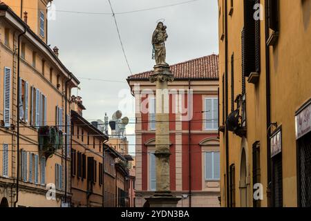 La Colonna della Beata Vergine del Carmine, créée par le sculpteur Andrea Ferreri en 1705, porte une statue en pierre de la Vierge Marie avec l’Enfant Jésus. Derrière elle, des antennes modernes en corne pour les communications mobiles s’élèvent au-dessus des toits de la Via Guglielmo Oberdan. Via Guglielmo Oberdan, Bologne, Émilie-Romagne, Italie. Banque D'Images