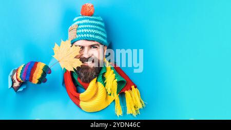 Homme barbu souriant avec feuille d'érable d'automne regardant à travers le trou dans le papier. Beau mâle en chapeau de laine, écharpe colorée et gants. Soldes d'automne et Banque D'Images