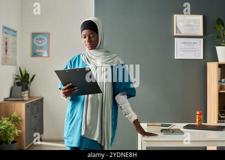 Professionnel de la santé musulman portant des gommages et debout dans un cadre de bureau moderne, examinant des cartes, avec des certificats et des diplômes sur le mur et divers articles de bureau autour Banque D'Images