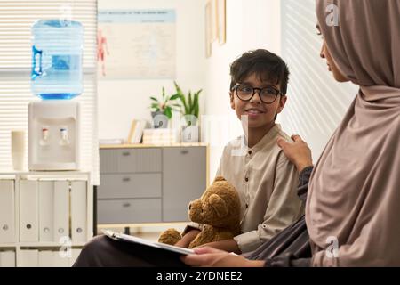 Garçon tenant un ours en peluche et souriant tout en étant assis en face d'une femme musulmane en hijab. Tous deux engagés dans la conversation dans un environnement de bureau moderne Banque D'Images