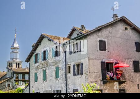 Un couple se détendent sur leur balcon tandis que le dôme d'oignon de l'église Saint Pancras s'élève derrière le hameau médiéval du XIVe siècle sur la rive française du lac Léman dans le petit village d'Yvoire, haute Savoie, France. Banque D'Images