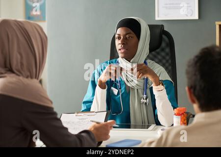 Médecin musulman en hijab assis en face du patient tout en écoutant attentivement dans la salle de consultation moderne en se concentrant sur les soins aux patients Banque D'Images