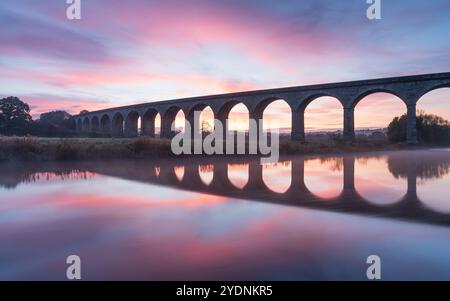 Un lever de soleil d'automne vif encadre les arches distinctives du viaduc d'Arthington, reflétées dans la rivière Wharfe par un matin d'octobre froid mais calme. Banque D'Images