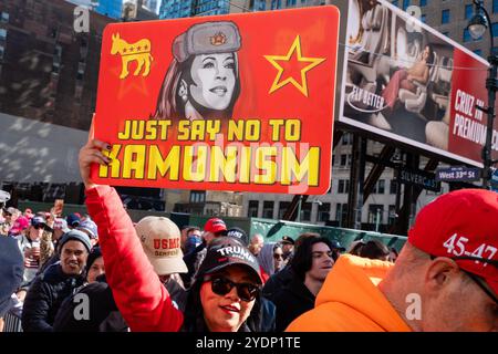 New York, NY, États-Unis. 27 octobre 2024. Des foules de partisans de Donald Trump, dont beaucoup portent des casquettes et des chemises de marque MAGA ou Trump, se rassemblent devant Madison Square Garden avant un rassemblement en soirée. Une femme dans la foule traversant la 8ème Avenue tient haut une pancarte avec une illustration de Kamala Harris dans une casquette de style russe avec des rabats d'oreille, en lisant "juste dire non au kamunisme." Crédit : Ed Lefkowicz/Alamy Live News Banque D'Images